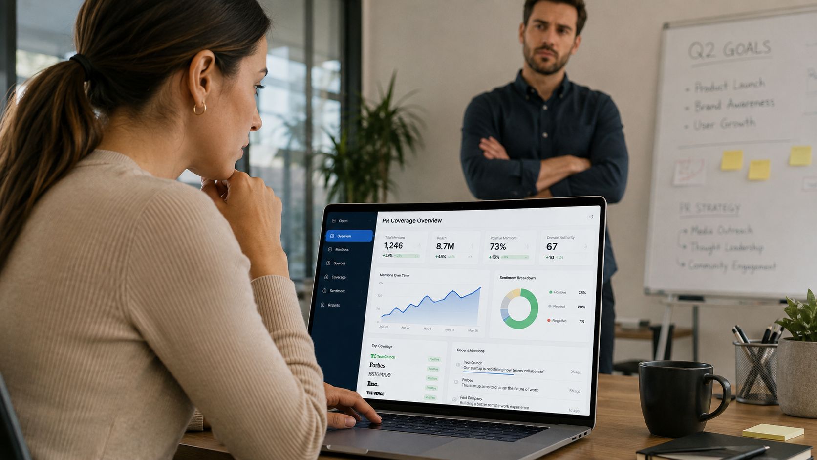 A professional woman analyzes PR coverage metrics on her laptop screen while a colleague stands behind her.